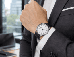 Detalhe do pulso de homem com terno cinza-escuro e camisa branca, relógio social Seculus de pulseira de couro preto com mostrador branco, ambiente de escritório com luz natural