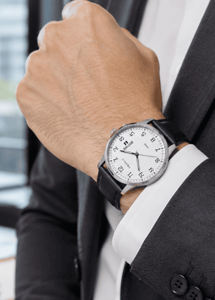 Detalhe do pulso de homem com terno cinza-escuro e camisa branca, relógio social Seculus de pulseira de couro preto com mostrador branco, ambiente de escritório com luz natural