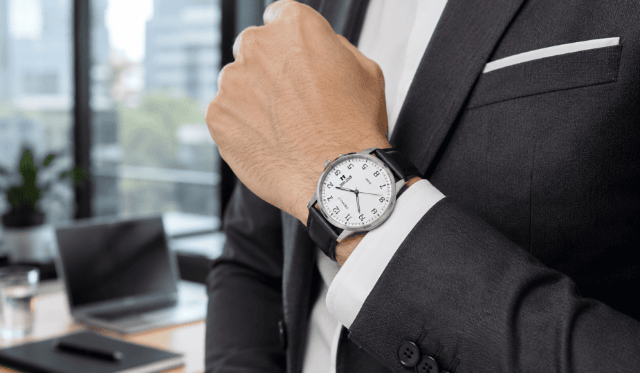 Detalhe do pulso de homem com terno cinza-escuro e camisa branca, relógio social Seculus de pulseira de couro preto com mostrador branco, ambiente de escritório com luz natural