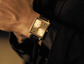 Close em pulso masculino usando relógio Seculus quadrado dourado com pulseira de couro sobre blazer escuro, destacando as linhas retas da caixa em contraste com a curvatura natural do pulso em estilo editorial sofisticado.