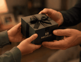 Mãos de um filho entregando uma caixa de presente Seculus ao pai, com foco no gesto de entrega. Ao fundo, ambiente desfocado com iluminação quente, transmitindo um clima familiar, acolhedor e afetuoso.