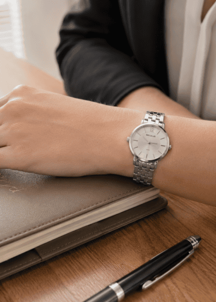 Mulher usando relógio feminino Seculus prata com pulseira de aço, mão apoiada sobre agenda de couro em mesa de escritório, blazer escuro ao fundo e iluminação natural lateral.