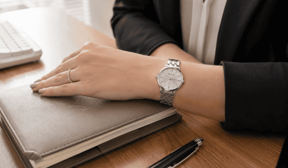 Mulher usando relógio feminino Seculus prata com pulseira de aço, mão apoiada sobre agenda de couro em mesa de escritório, blazer escuro ao fundo e iluminação natural lateral.