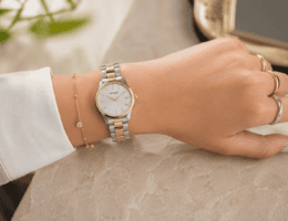 Pulso feminino com relógio bicolor Seculus de pulseira em aço prata e dourado, mostrador claro e caixa delicada, combinado com anéis em prata e ouro sobre fundo suave com iluminação natural.
