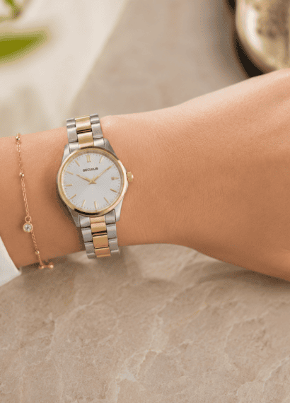 Pulso feminino com relógio bicolor Seculus de pulseira em aço prata e dourado, mostrador claro e caixa delicada, combinado com anéis em prata e ouro sobre fundo suave com iluminação natural.