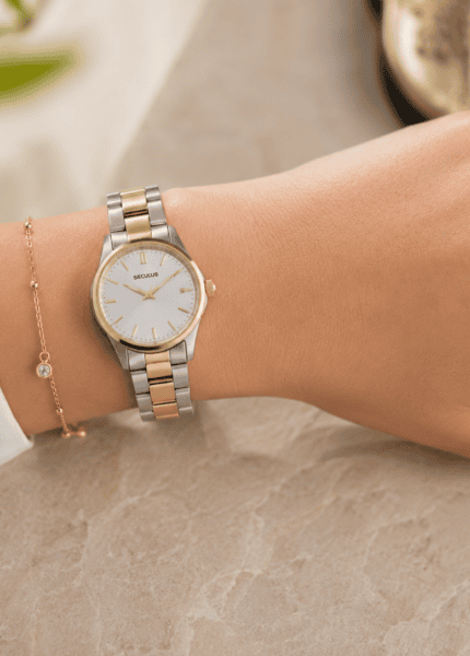 Pulso feminino com relógio bicolor Seculus de pulseira em aço prata e dourado, mostrador claro e caixa delicada, combinado com anéis em prata e ouro sobre fundo suave com iluminação natural.