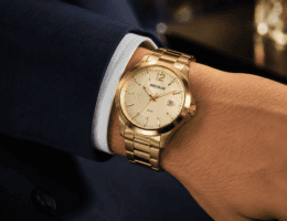 Pulso masculino usando relógio dourado Seculus com pulseira de aço sobre manga de terno azul-marinho, em close com fundo desfocado e iluminação suave.