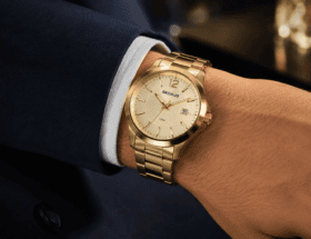 Pulso masculino usando relógio dourado Seculus com pulseira de aço sobre manga de terno azul-marinho, em close com fundo desfocado e iluminação suave.