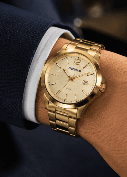 Pulso masculino usando relógio dourado Seculus com pulseira de aço sobre manga de terno azul-marinho, em close com fundo desfocado e iluminação suave.