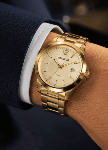 Pulso masculino usando relógio dourado Seculus com pulseira de aço sobre manga de terno azul-marinho, em close com fundo desfocado e iluminação suave.