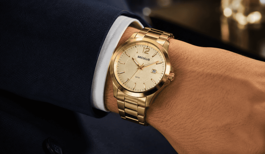 Pulso masculino usando relógio dourado Seculus com pulseira de aço sobre manga de terno azul-marinho, em close com fundo desfocado e iluminação suave.