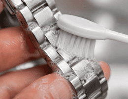 Escova de dentes extramacia limpando suavemente os elos de uma pulseira de aço com espuma de sabão.