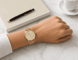 Relógio Seculus feminino dourado com pulseira milanesa no pulso, apoiado sobre mesa de mármore branco ao lado de caderno, caneta e xícara de porcelana.