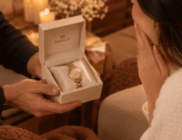 Mãos masculinas entregam caixa de presente Seculus aberta com relógio feminino dourado, enquanto mulher recebe o presente em clima romântico e afetuoso.