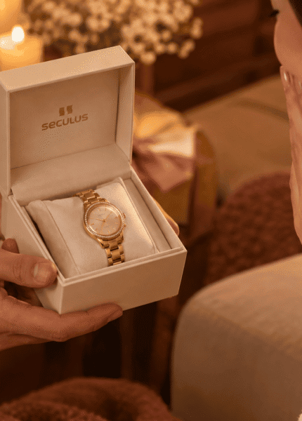 Mãos masculinas entregam caixa de presente Seculus aberta com relógio feminino dourado, enquanto mulher recebe o presente em clima romântico e afetuoso.