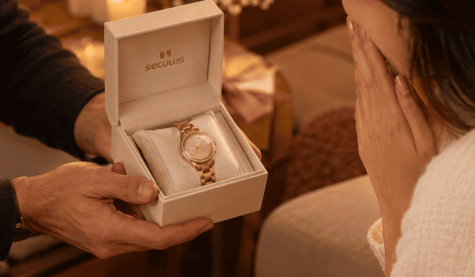 Mãos masculinas entregam caixa de presente Seculus aberta com relógio feminino dourado, enquanto mulher recebe o presente em clima romântico e afetuoso.