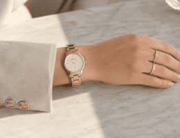 Relógio feminino Seculus dourado delicado no pulso, com anéis finos, blazer creme e mão apoiada sobre mármore claro.