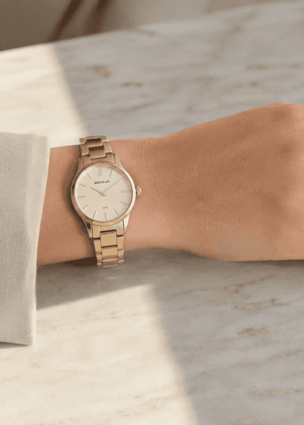 Relógio feminino Seculus dourado delicado no pulso, com anéis finos, blazer creme e mão apoiada sobre mármore claro.