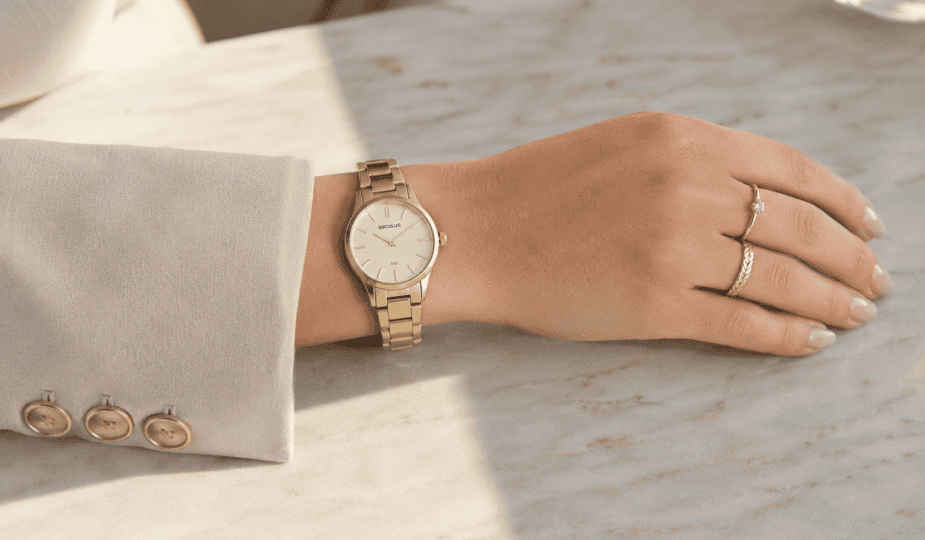 Relógio feminino Seculus dourado delicado no pulso, com anéis finos, blazer creme e mão apoiada sobre mármore claro.