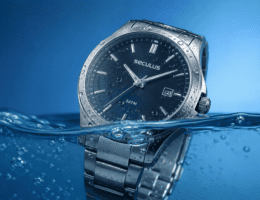 Relógio Seculus prata parcialmente submerso em água cristalina, com gotas no vidro e fundo azul degradê.