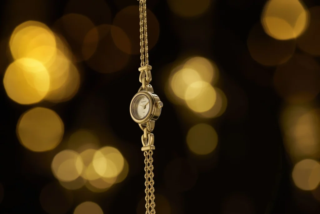 Close do Relógio Seculus Eternity Dourado Slim posicionado verticalmente, destacando sua caixa pequena e delicada, a pulseira fina em formato de corrente dourada e o design minimalista contra um fundo escuro com luzes bokeh douradas.