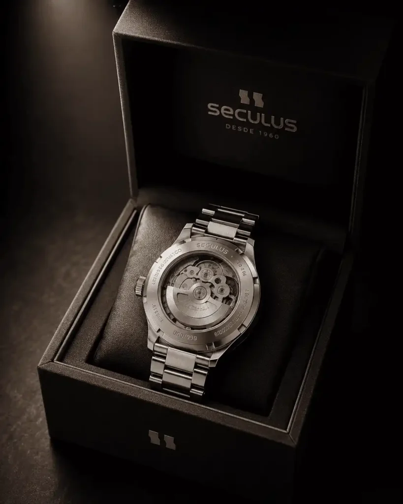 Relógio automático Seculus masculino em aço prata com fundo de vidro exibindo o mecanismo interno, acomodado em caixa de presente premium da marca sobre fundo escuro com iluminação direcionada.