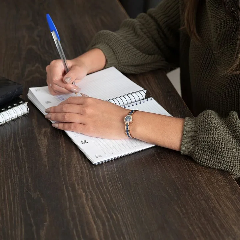Mulher escrevendo em uma agenda enquanto usa Relógio Feminino Seculus Cerâmica Cristais Rosé, caixa dourada e pulseira bicolor em azul e dourado, destacando delicadeza e elegância para a rotina.
