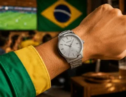 Pulso masculino usando relógio Seculus prateado com pulseira de aço, combinado com camisa verde e amarela. Ao fundo, ambiente desfocado de sala ou bar com televisão exibindo partida de futebol e bandeira do Brasil, em clima vibrante e iluminação natural.