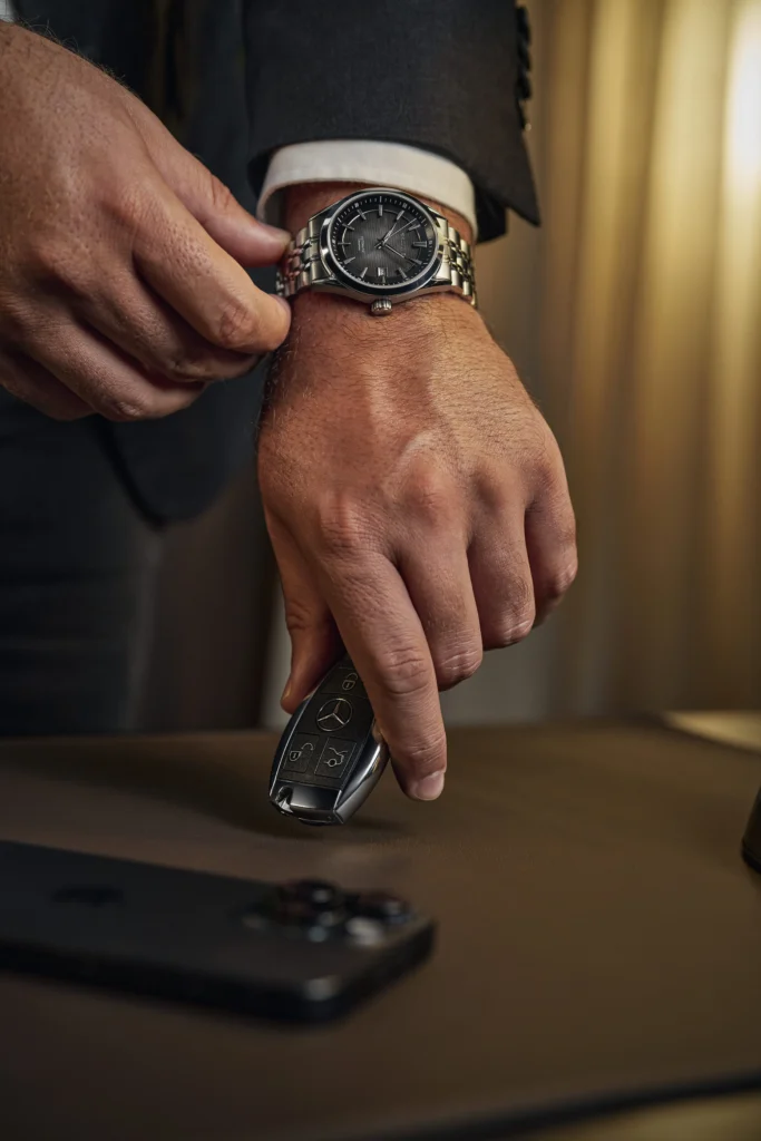  Homem ajustando um relógio cronógrafo masculino de aço inoxidável no pulso enquanto segura uma chave automotiva de luxo, em ambiente sofisticado com iluminação quente e elegante.