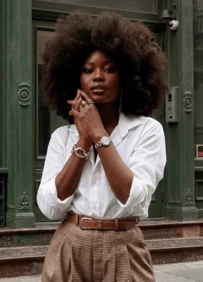 Mulher usando relógio feminino de aço prateado com mostrador claro, combinado com camisa branca e calça de alfaiataria bege, em cenário urbano de arquitetura clássica.