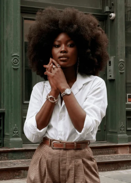 Mulher usando relógio feminino de aço prateado com mostrador claro, combinado com camisa branca e calça de alfaiataria bege, em cenário urbano de arquitetura clássica.