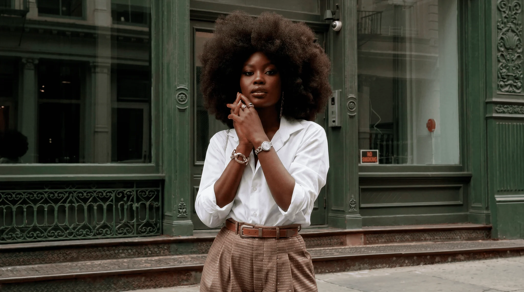 Mulher usando relógio feminino de aço prateado com mostrador claro, combinado com camisa branca e calça de alfaiataria bege, em cenário urbano de arquitetura clássica.