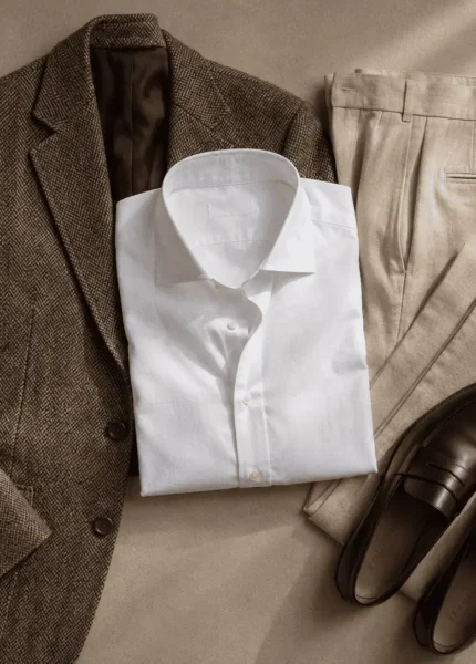 Composição de roupas old money sobre superfície neutra: blazer de tweed, calça de linho bege, camisa de tricoline branca e mocassim de couro liso