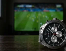 Relógio de pulso prateado da marca Seculus com mostrador preto e cronógrafo, posicionado em cima de uma mesa de madeira escura. Ao fundo, uma televisão desfocada exibe uma partida de futebol em um gramado verde.