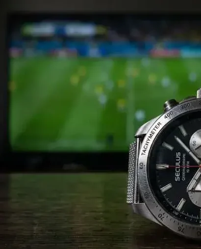 Relógio de pulso prateado da marca Seculus com mostrador preto e cronógrafo, posicionado em cima de uma mesa de madeira escura. Ao fundo, uma televisão desfocada exibe uma partida de futebol em um gramado verde.