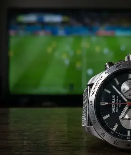 Relógio de pulso prateado da marca Seculus com mostrador preto e cronógrafo, posicionado em cima de uma mesa de madeira escura. Ao fundo, uma televisão desfocada exibe uma partida de futebol em um gramado verde.