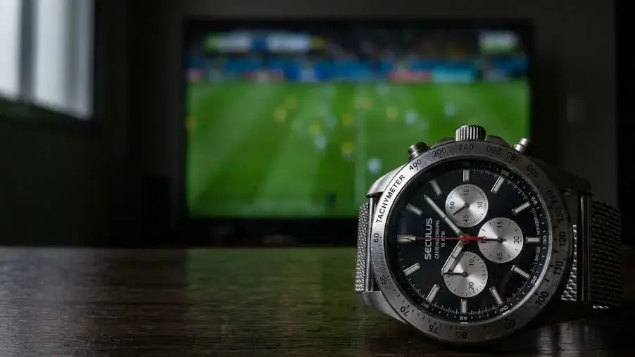 Relógio de pulso prateado da marca Seculus com mostrador preto e cronógrafo, posicionado em cima de uma mesa de madeira escura. Ao fundo, uma televisão desfocada exibe uma partida de futebol em um gramado verde.