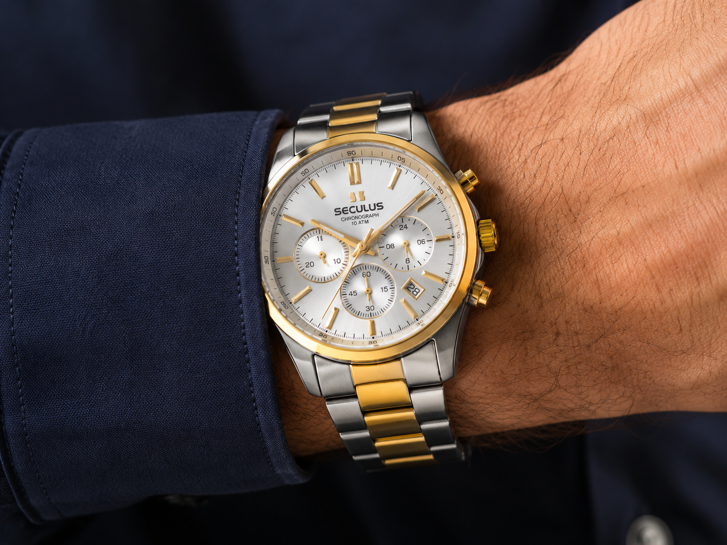Close do relógio masculino cronógrafo Seculus com pulseira bicolor em aço prata e dourado no pulso sobre manga de camisa social azul-marinho, destacando os submostradores e o acabamento metálico sob luz lateral.