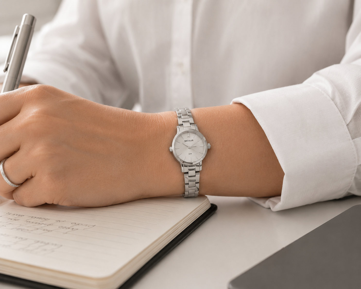 Close no pulso feminino com relógio Seculus prata de caixa mini e pulseira de aço, camisa social branca com manga dobrada e mão segurando caneta sobre caderno em ambiente executivo clean.