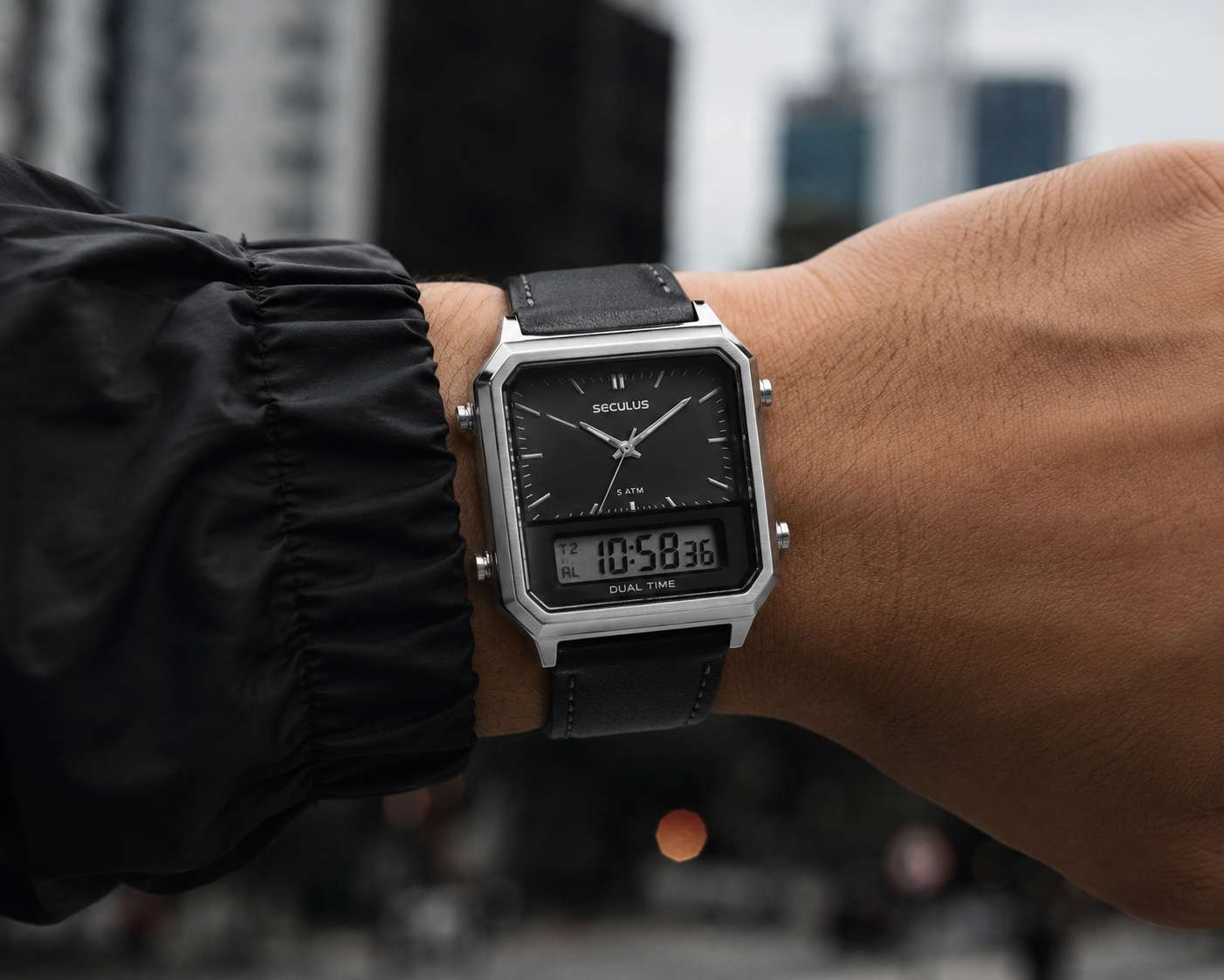Homem usando relógio masculino Seculus anadigi de caixa quadrada prateada e pulseira de couro preta no pulso, sobre manga de jaqueta corta-vento preta, com fundo urbano desfocado e foco no mostrador analógico e digital.