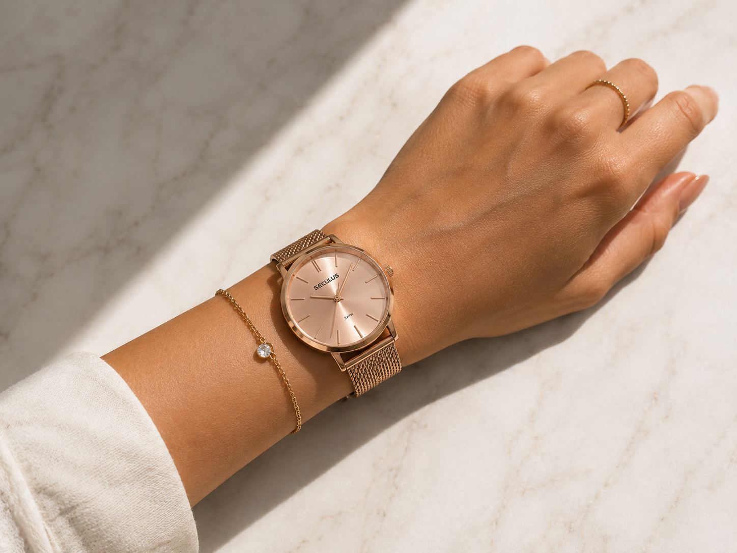 Mão feminina com anel dourado fino e pulseira delicada, relógio Seculus de pulseira milanesa rosê no pulso, fundo de mármore claro, luz natural lateral