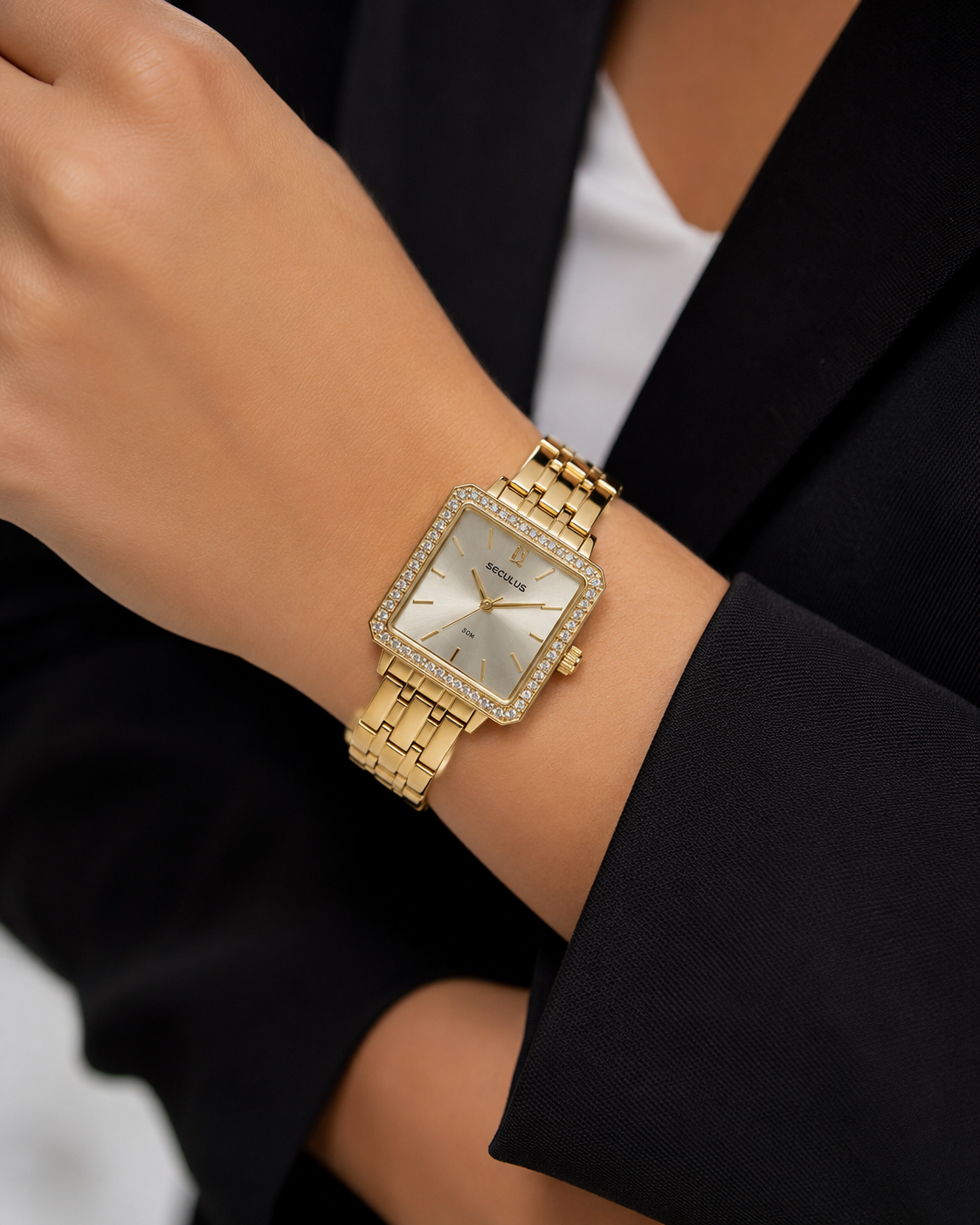 Mulher usando relógio feminino Seculus analógico dourado com caixa quadrada, pedras cravejadas no aro e pulseira de aço dourada, combinado com blazer escuro em estilo elegante e corporativo.