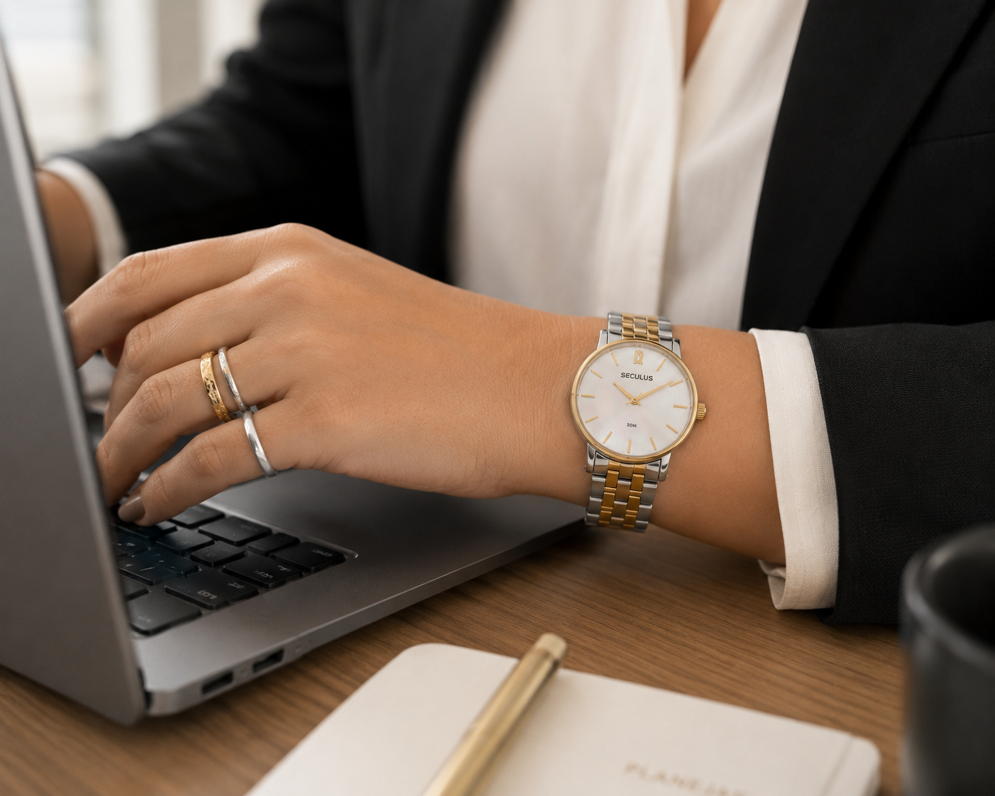 Mulher usando relógio feminino Seculus com mostrador em madrepérola, pulseira bicolor prata e dourado e anéis mistos, com a mão sobre um laptop em mesa de escritório, em ambiente profissional e elegante.