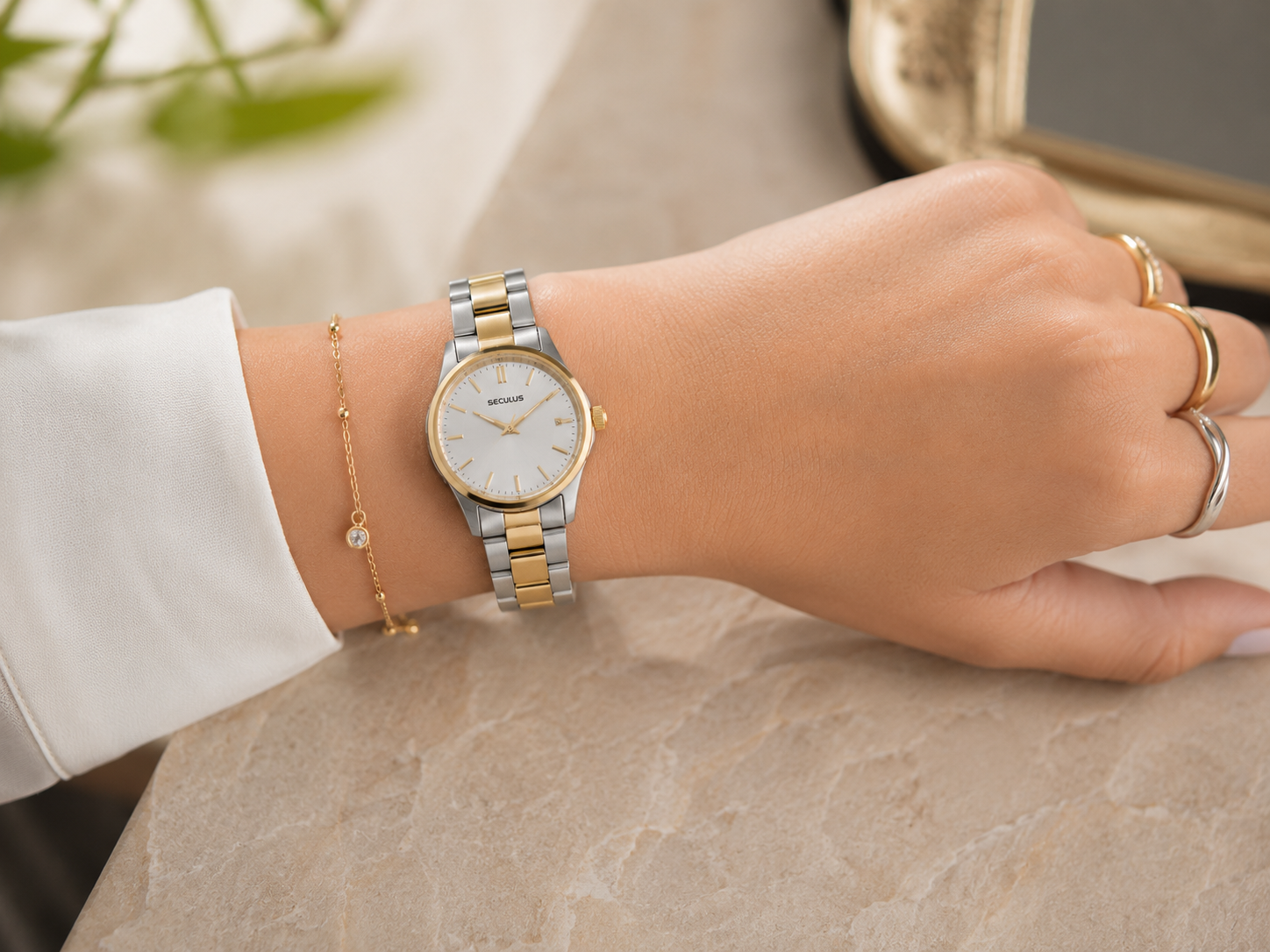 Pulso feminino com relógio bicolor Seculus de pulseira em aço prata e dourado, mostrador claro e caixa delicada, combinado com anéis em prata e ouro sobre fundo suave com iluminação natural.