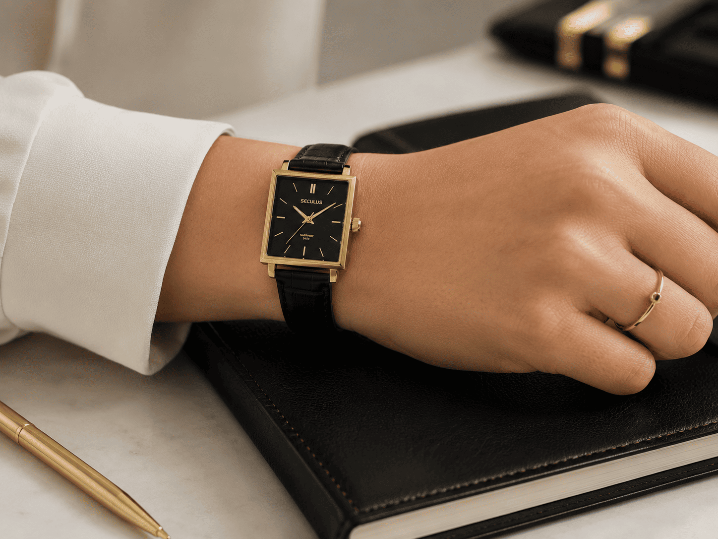Pulso feminino com Relógio Feminino Quadrado Minimalista Couro Preto Seculus ao lado de um anel fino dourado, apoiado sobre um caderno de couro preto, em composição clean e profissional.
