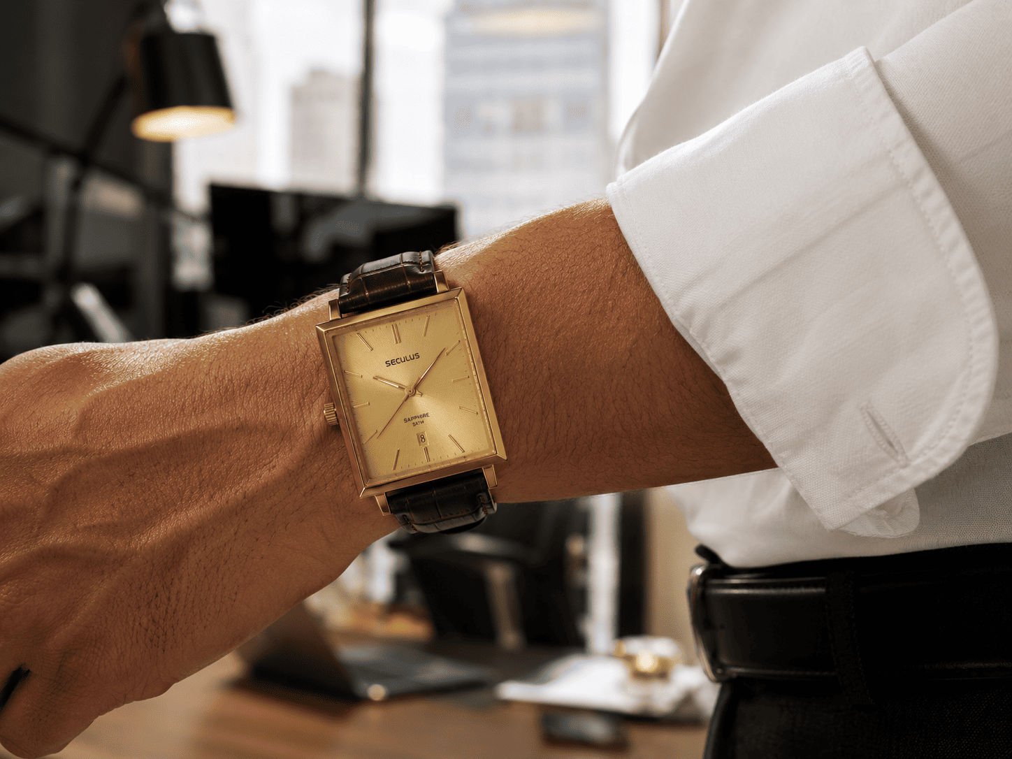 Pulso masculino com relógio quadrado dourado Seculus e pulseira de couro, usado com camisa social branca de mangas dobradas em escritório moderno desfocado, transmitindo um estilo corporativo elegante.
