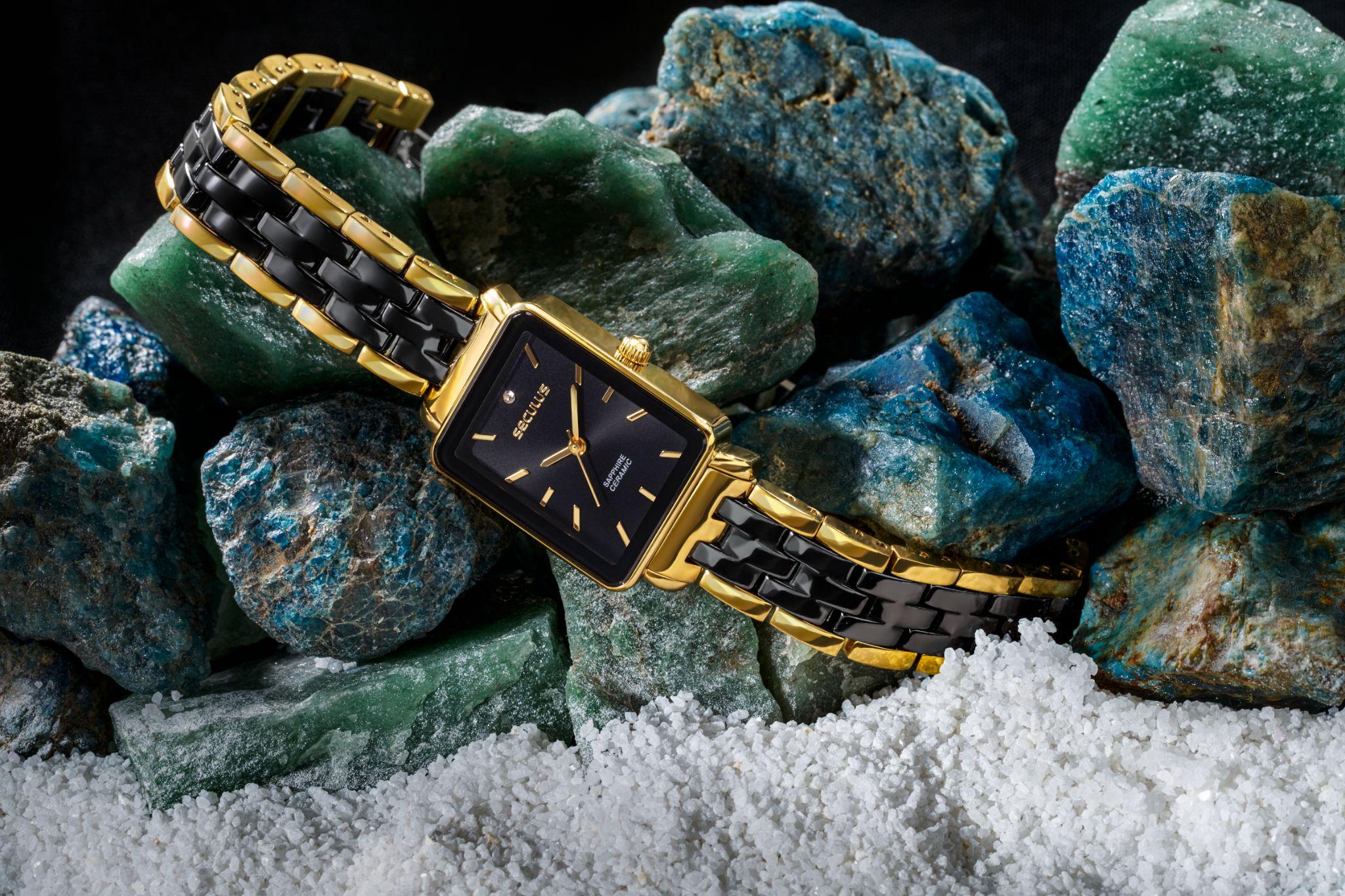 Relógio Feminino Cerâmica Quadrado Preto Dourado com caixa quadrada e mostrador preto, apoiado sobre pedras naturais em composição sofisticada, destacando a pulseira em cerâmica e aço dourado.