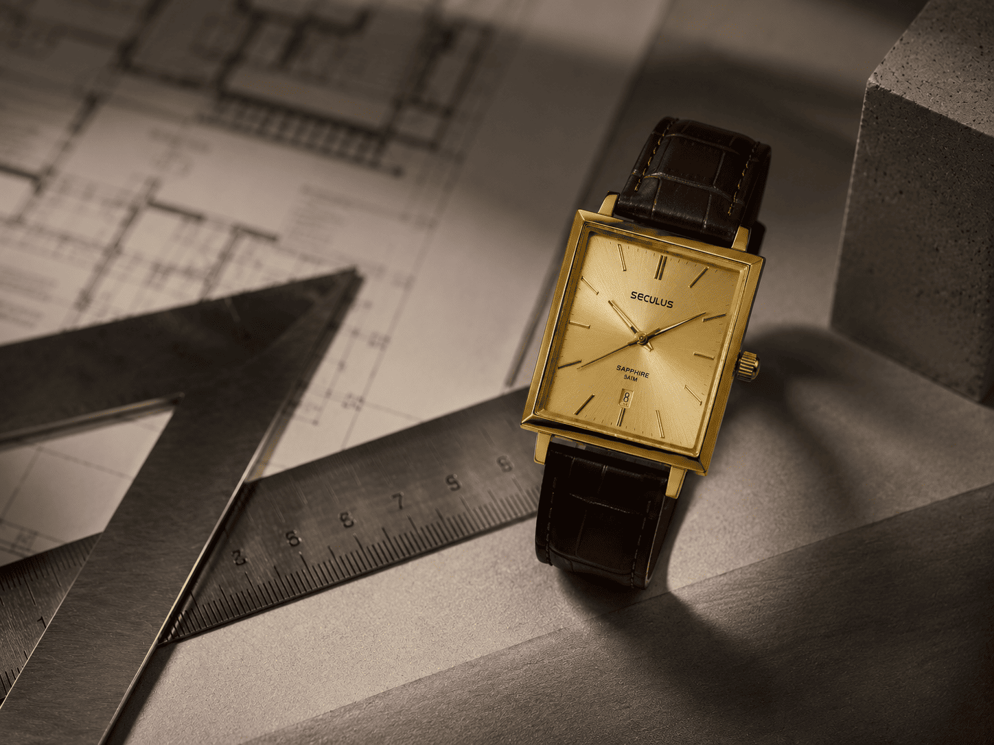 Relógio Seculus quadrado dourado com pulseira de couro ao lado de régua, esquadro e planta arquitetônica desfocada, em composição editorial que destaca o design geométrico e a conexão entre arquitetura e relojoaria.
