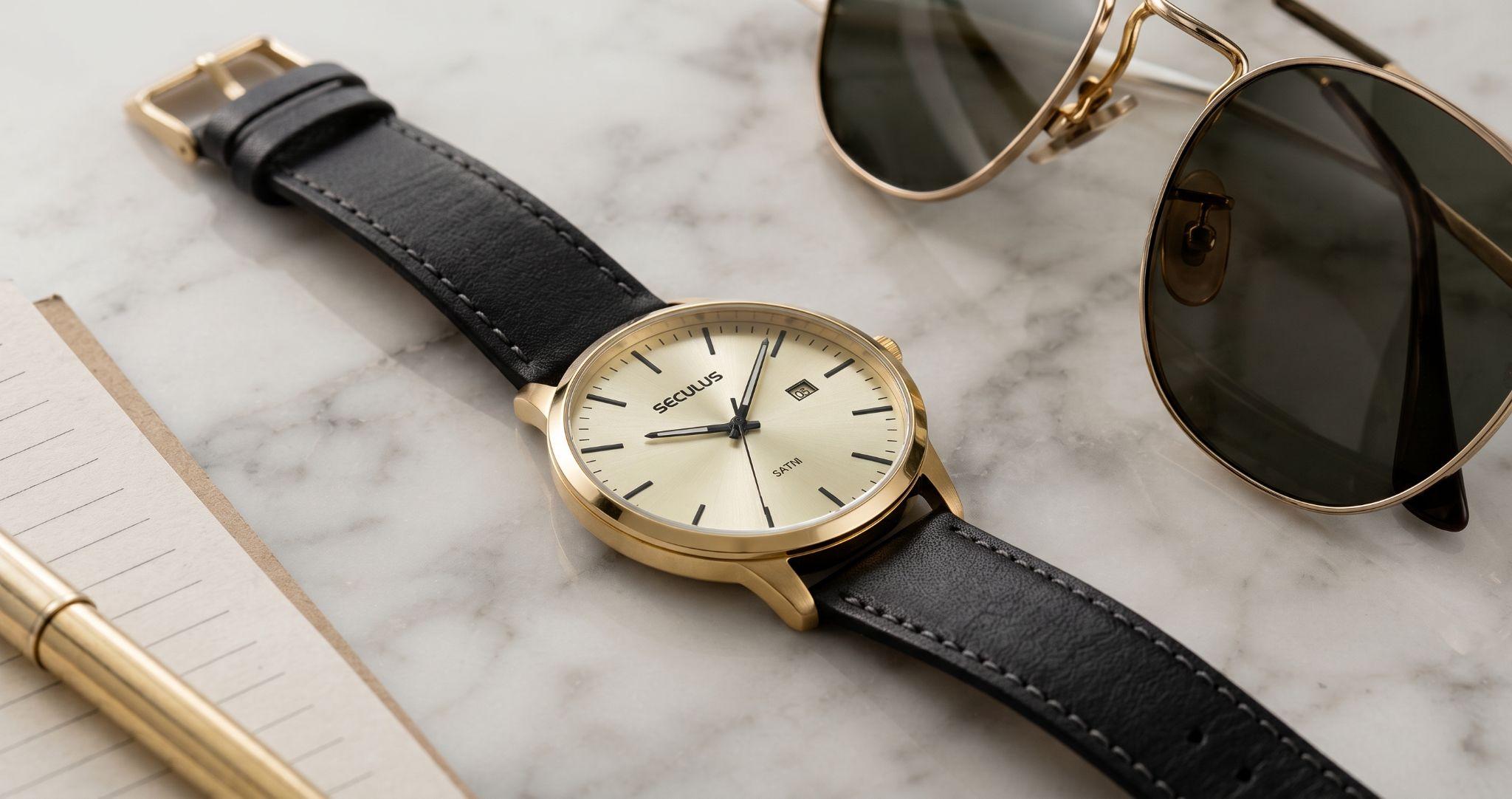 Relógio masculino Seculus slim dourado com caixa fina e pulseira de couro marrom com textura croco, apoiado sobre superfície de mármore branco, ao lado de óculos de sol de armação fina, em composição sofisticada e elegante.