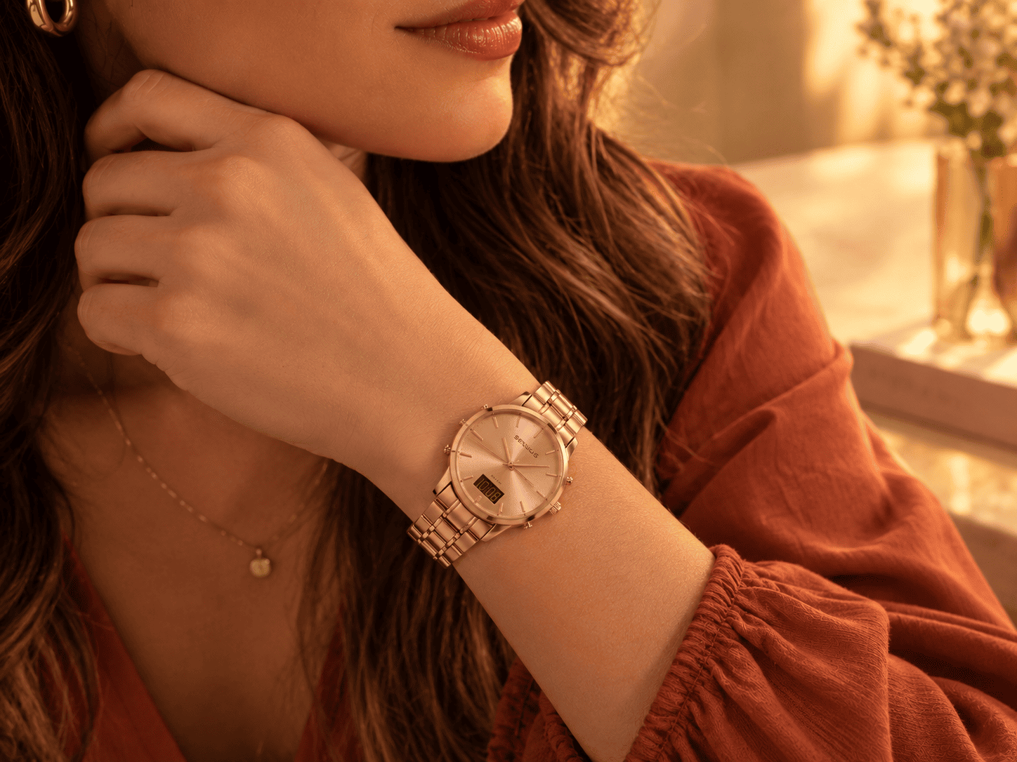 Relógio feminino rosé Seculus no pulso, combinado com vestido terracota e iluminado por luz dourada em ambiente acolhedor.